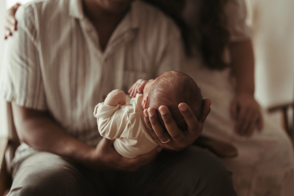Parents gently cradling their swaddled newborn baby together in soft warm indoor light.