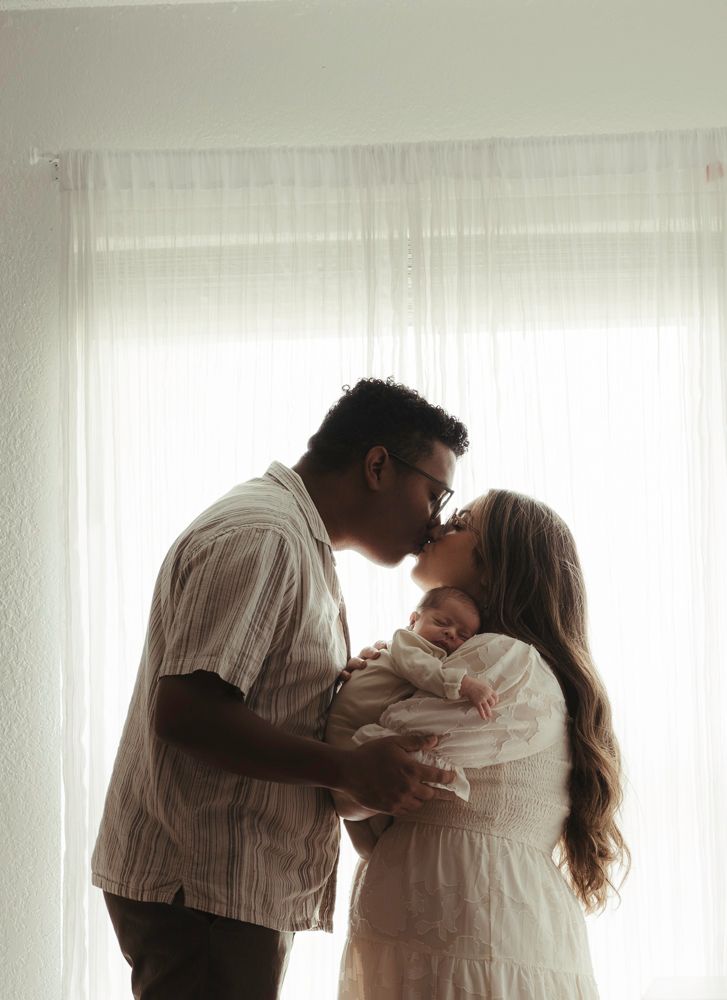 Couple sharing a tender kiss while holding their newborn baby in soft natural light by a window.