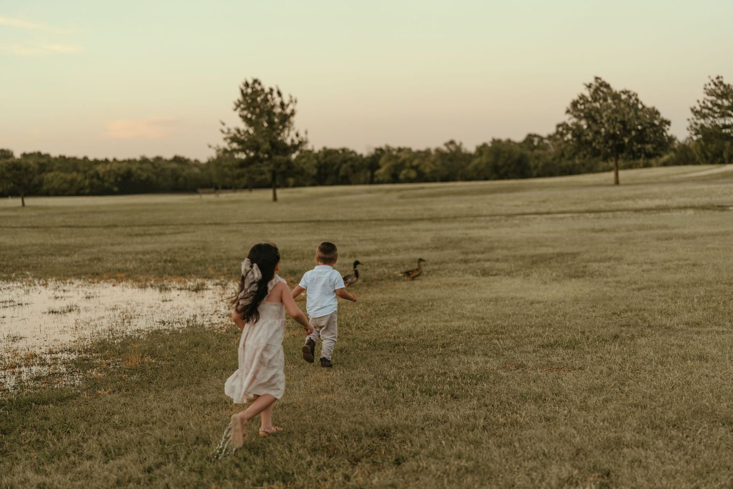 Two children run hand-in-hand across an open field at sunset near a small pond.