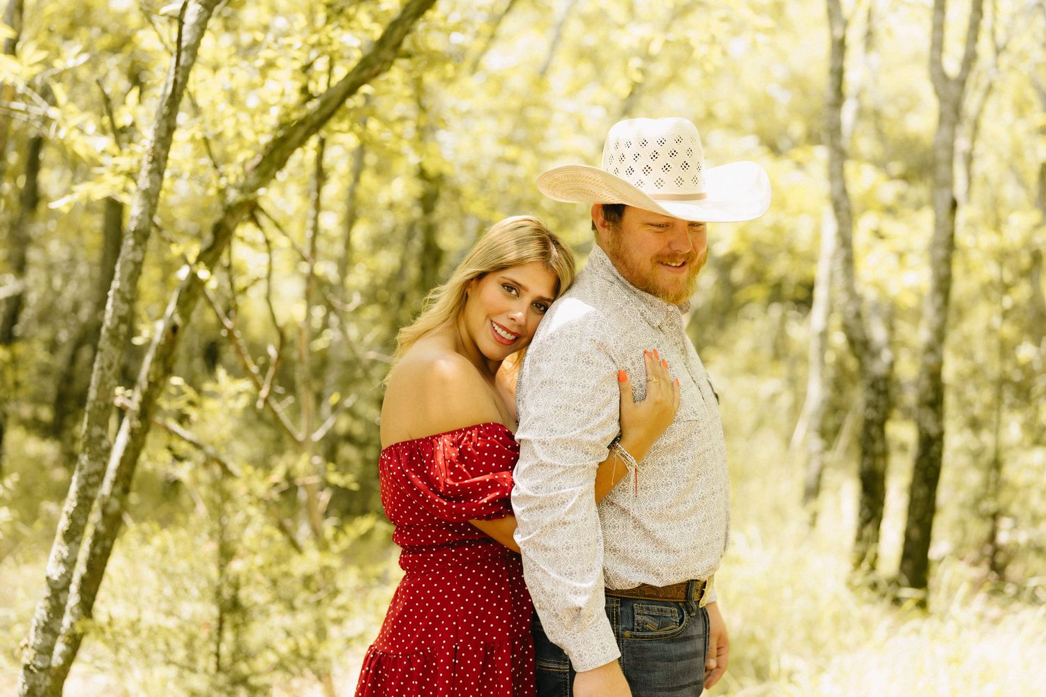 Couple posing outdoors in a forest, woman in red off-shoulder dress, man in white cowboy hat and plaid shirt.