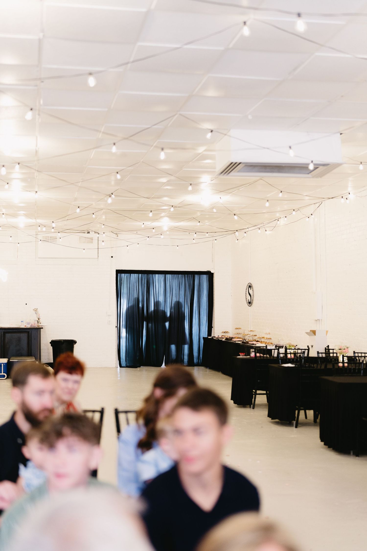 Elegant wedding reception hall with string lights on ceiling, black draped curtains, and guests seated at round tables.