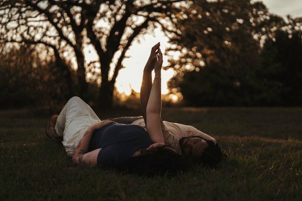 Couple lying on grass holding hands up toward sunset sky in romantic outdoor portrait.