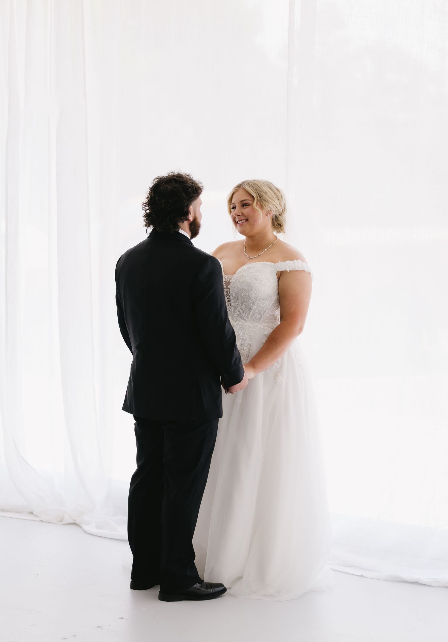 Bride in off-shoulder lace gown smiles at groom in black suit during elegant first look.