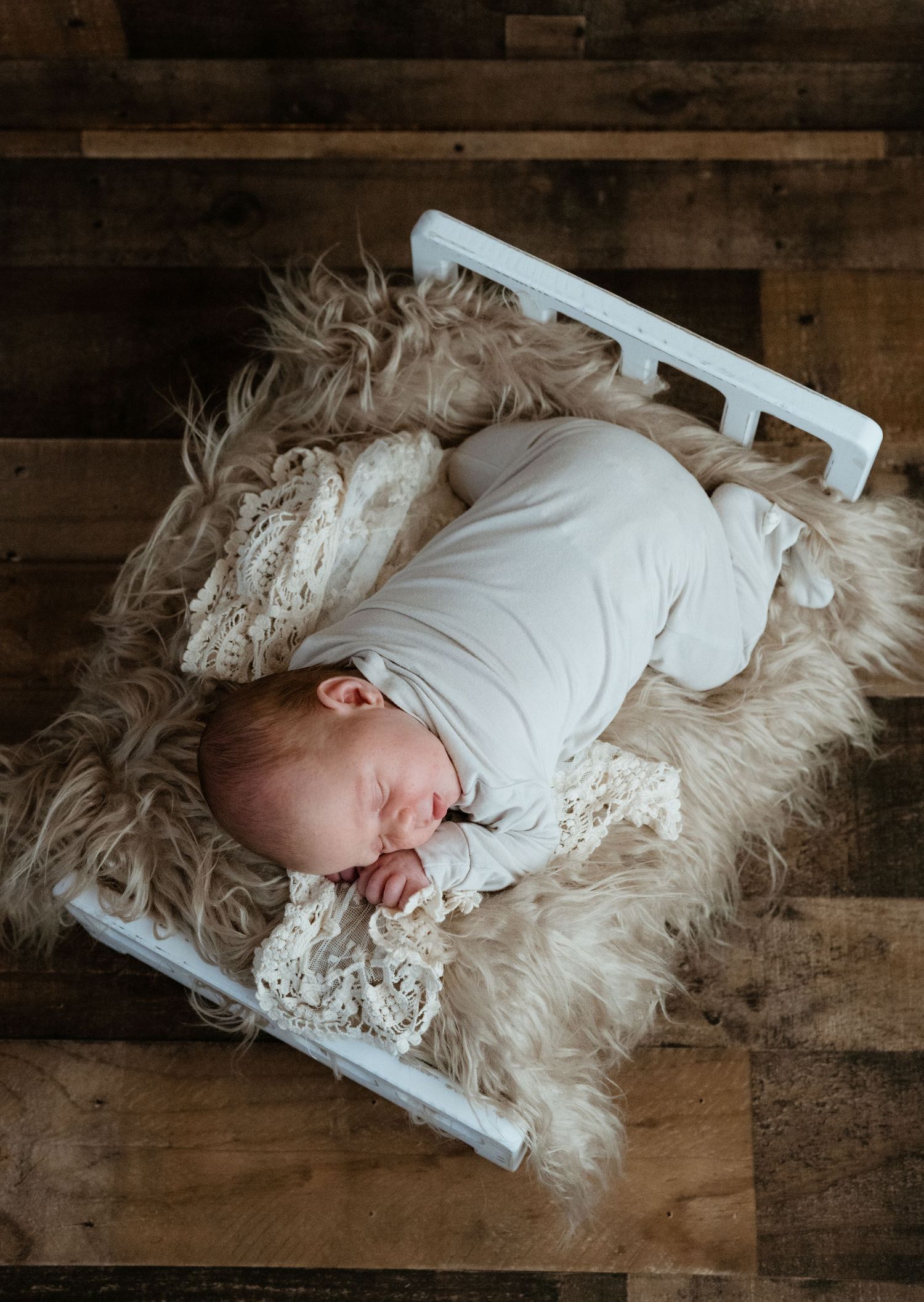 Peaceful newborn baby wrapped in white swaddle sleeping on tiny white bed with lace and fur on wood floor.