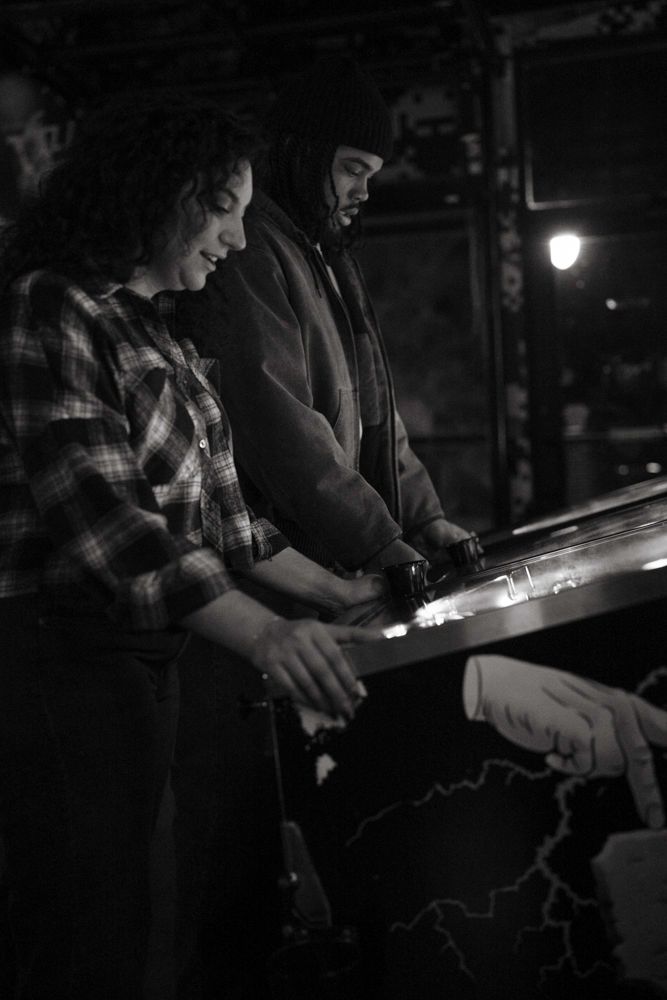Black and white photo of smiling woman in plaid shirt and bearded man playing pinball together in dark arcade.