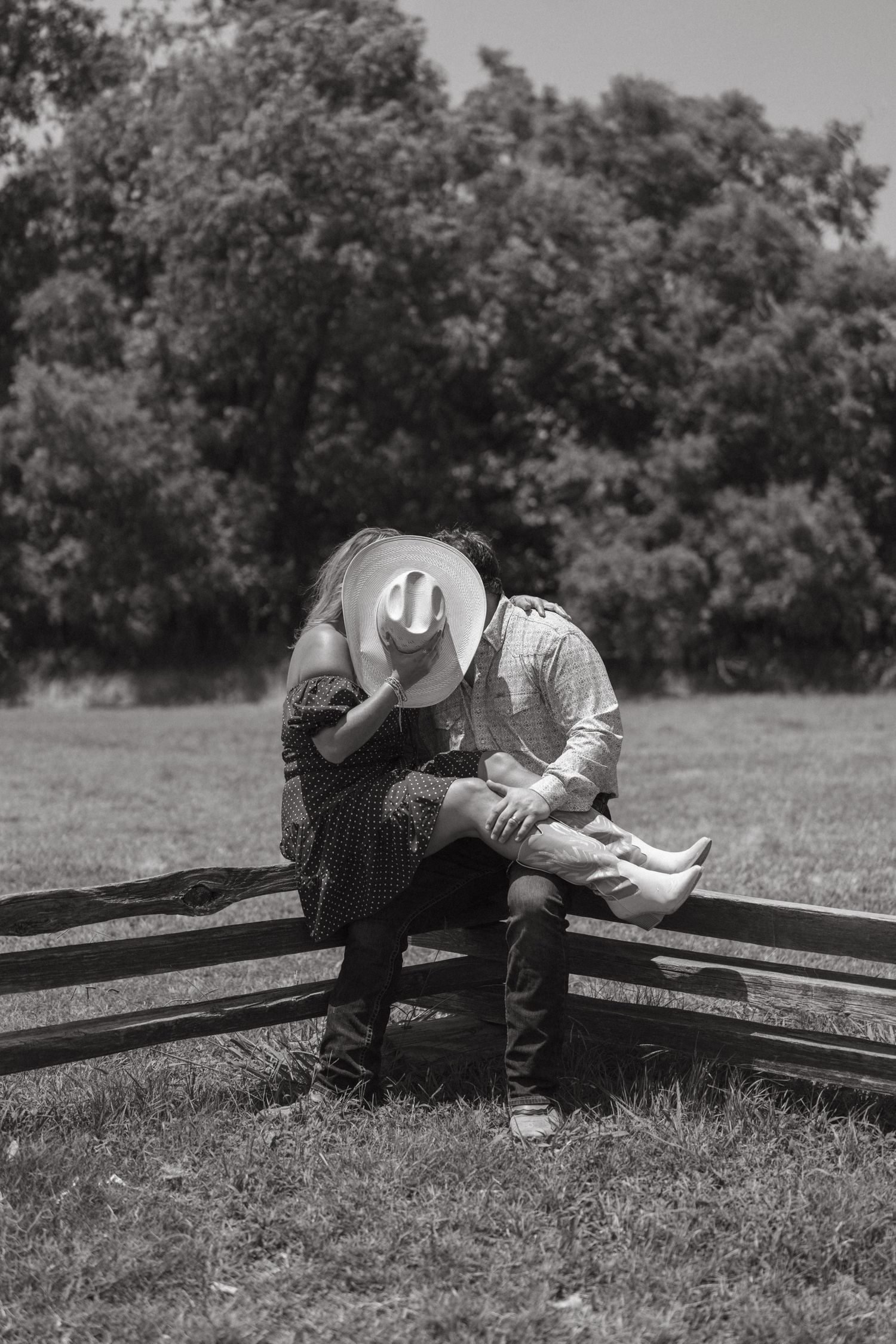 Black and white photo of a couple kissing behind a cowboy hat on a wooden fence in a rural field.