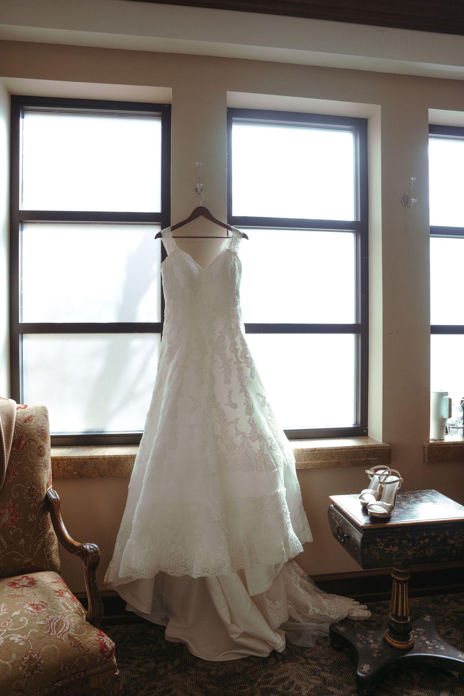 White lace A-line wedding dress hanging on wooden hanger in front of three windows with natural light.