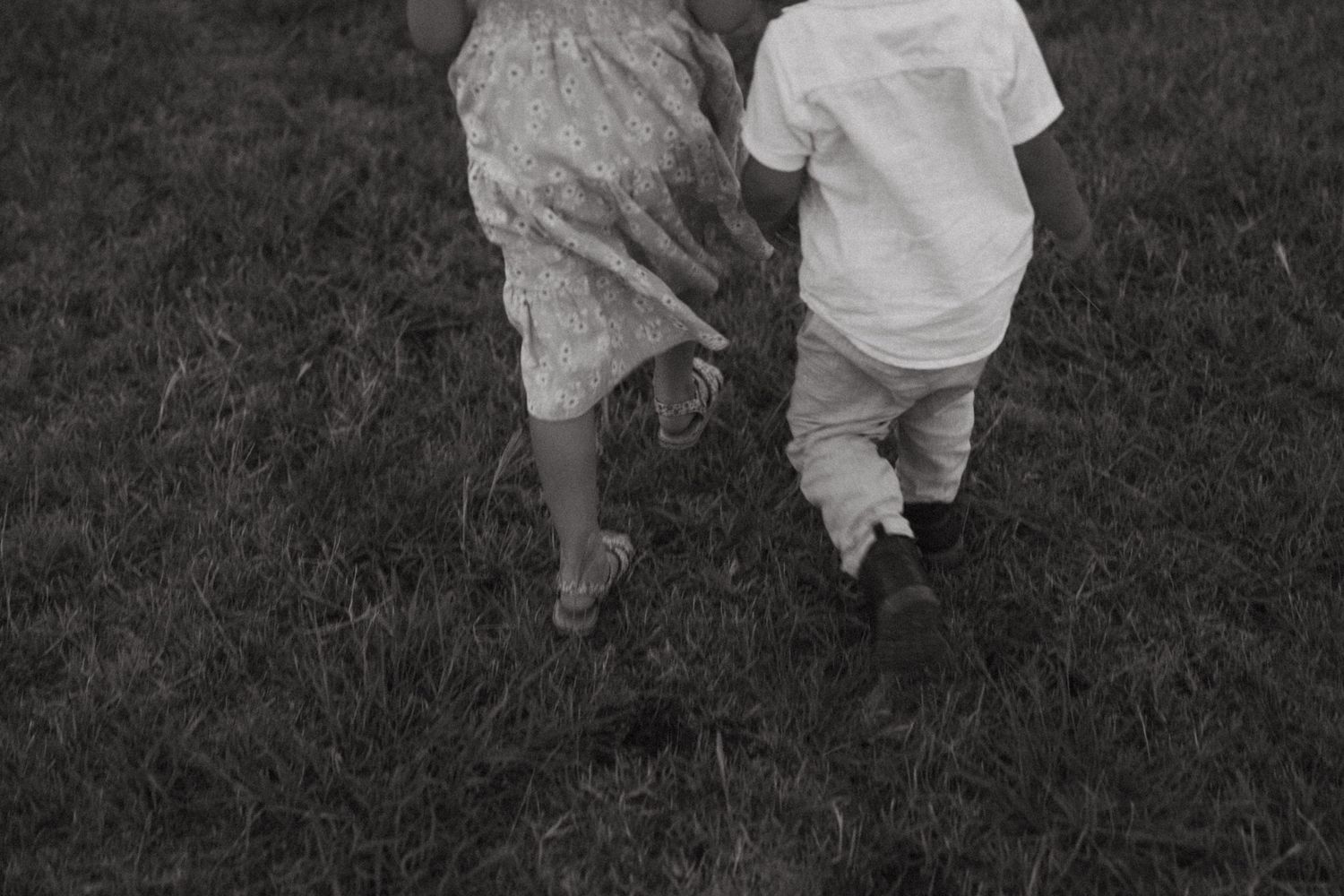 Black and white photo of two children holding hands while running through a grassy field.