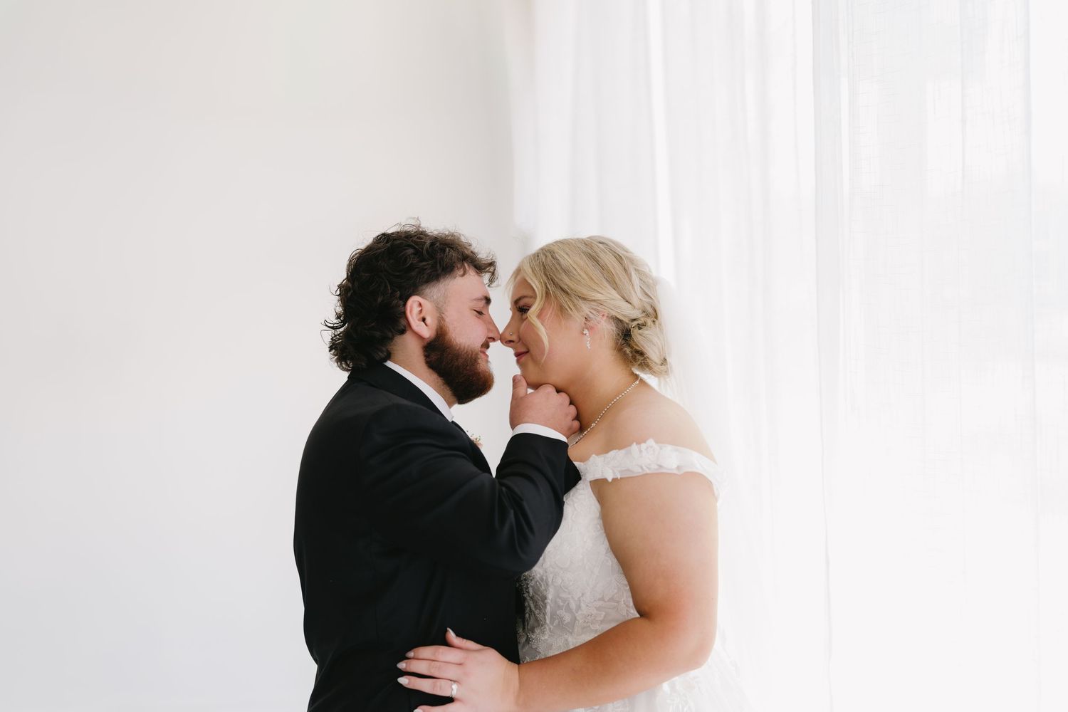 Bride and bearded groom share a tender embrace near sheer white curtains in soft light.