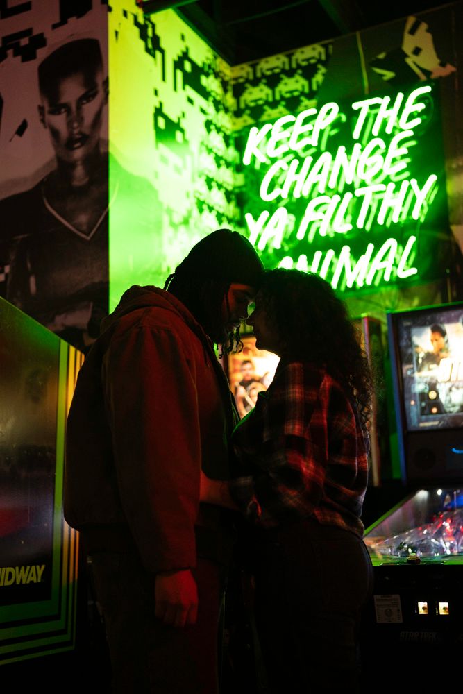 Couple kissing in front of a green neon sign reading Keep The Change Ya Filthy Animal in an arcade bar.