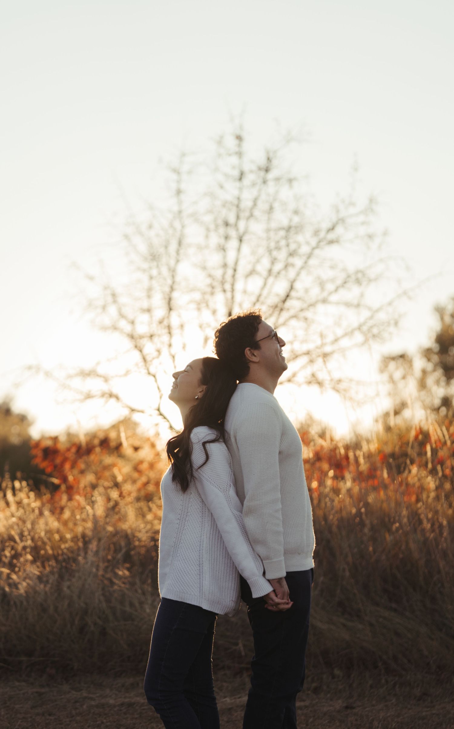 Engaged couple standing back to back holding hands in autumn field at golden hour.