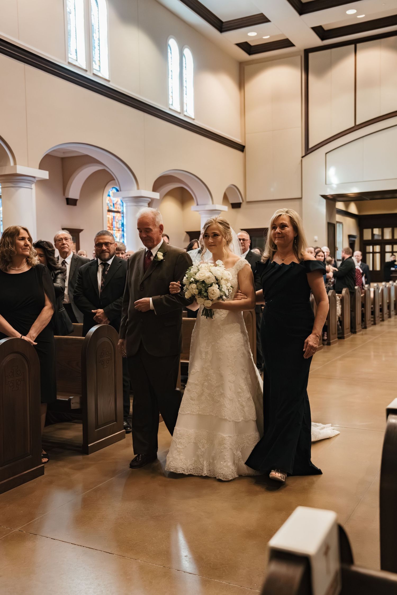 Bride in white lace gown with white bouquet walks down church aisle escorted by parents at formal wedding.