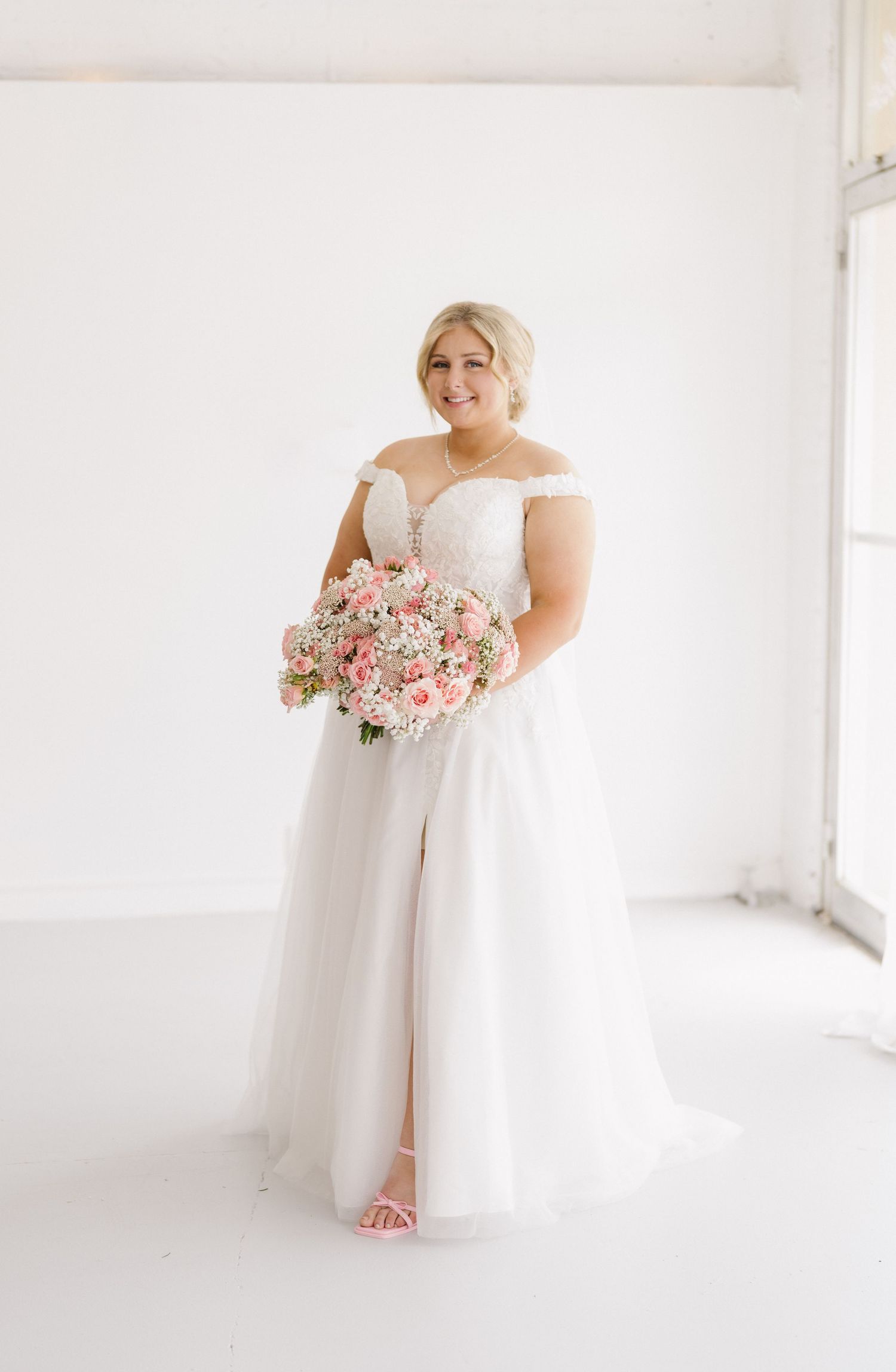 Radiant plus-size bride in off-shoulder tulle gown holds pink floral bouquet in bright studio.