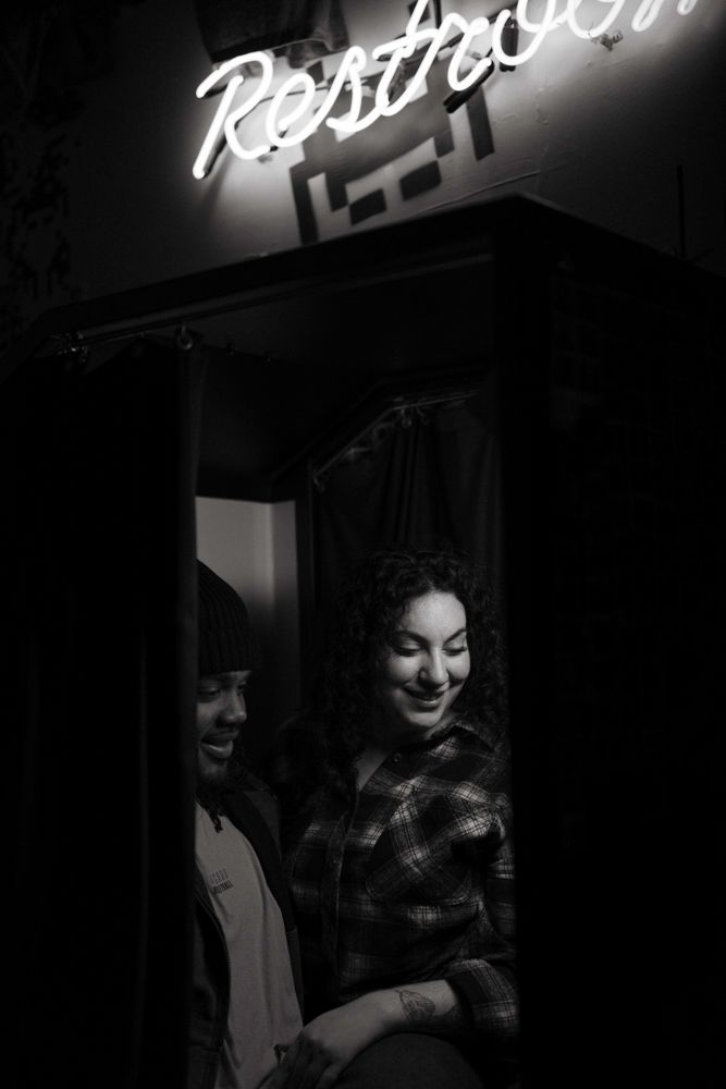 Black and white photo of a smiling couple standing beneath a glowing Restroom neon sign.