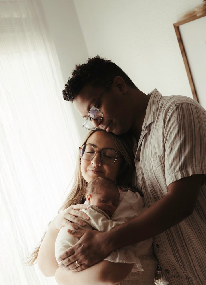 New parents with glasses lovingly holding their newborn baby by a sunlit window.