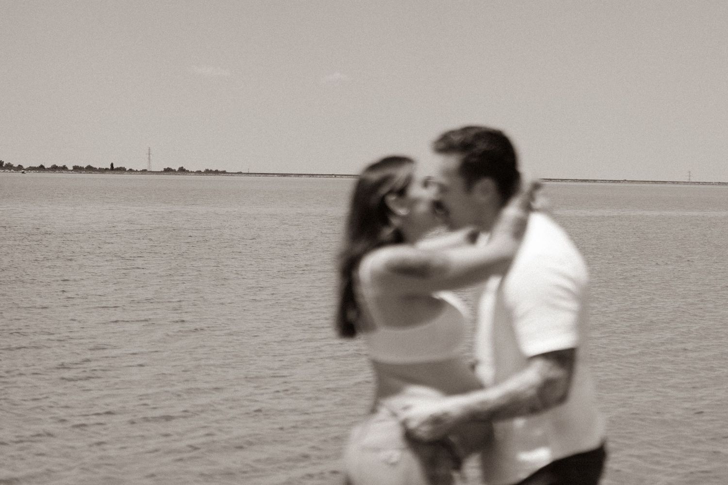 Vintage-style black and white photo of couple kissing by calm ocean waterfront.