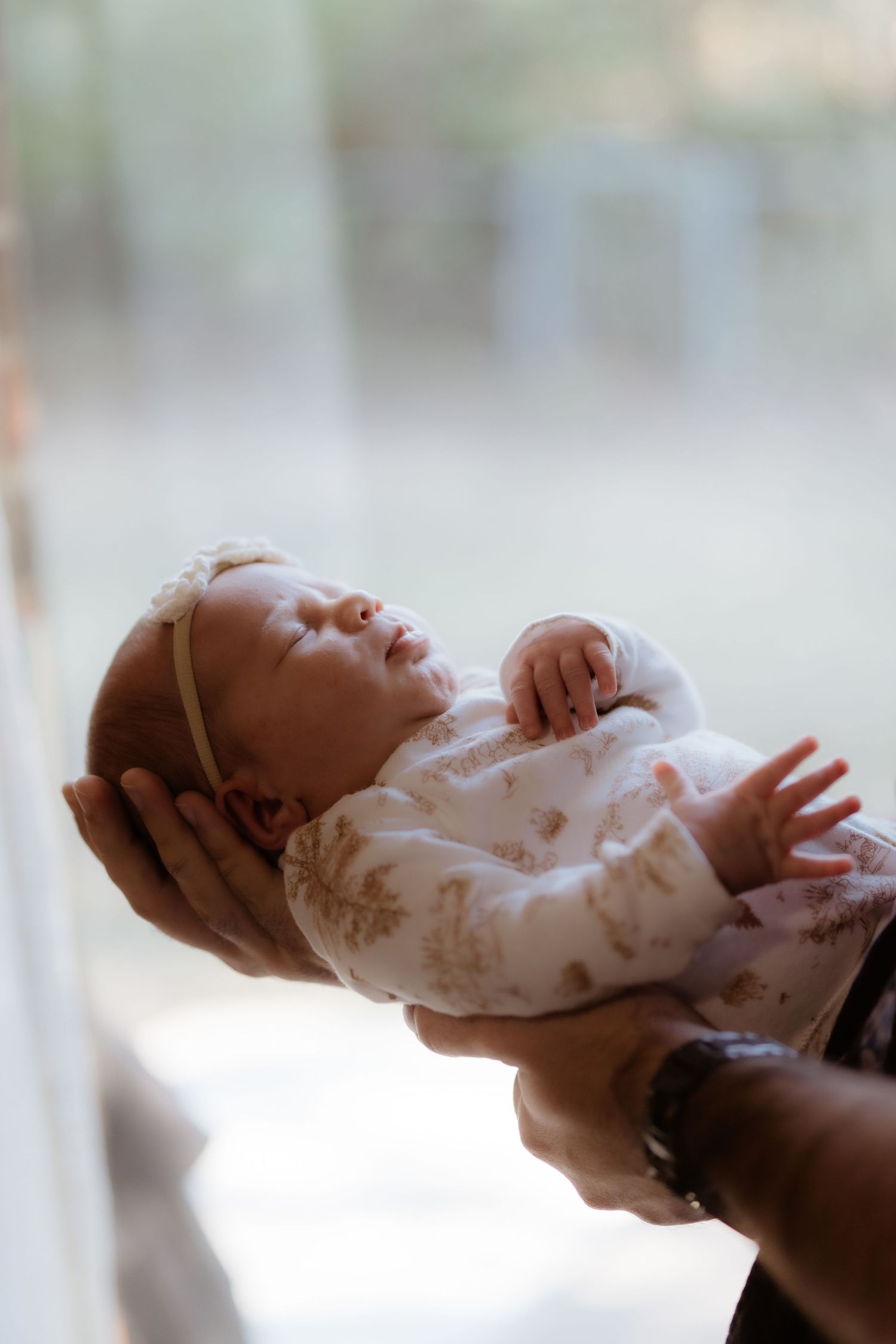 Newborn baby in floral onesie with bow headband gently cradled in parents hands near bright window.