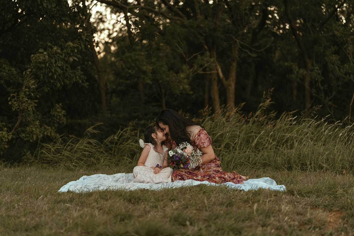 Two girls sit together on a white blanket in a meadow sharing a bouquet at golden hour.