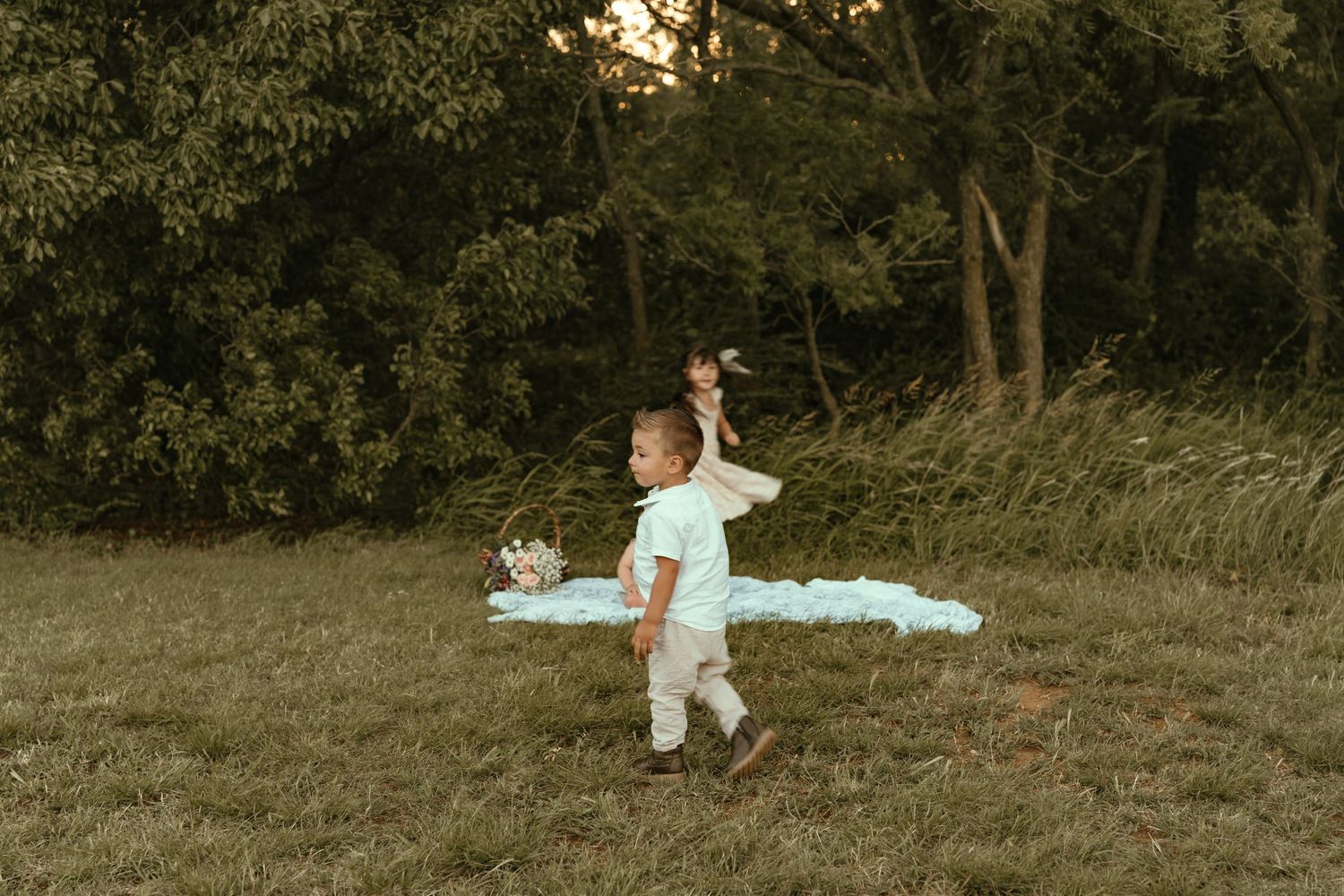 A toddler plays outdoors in a green meadow during golden hour, holding a toy near a picnic blanket.