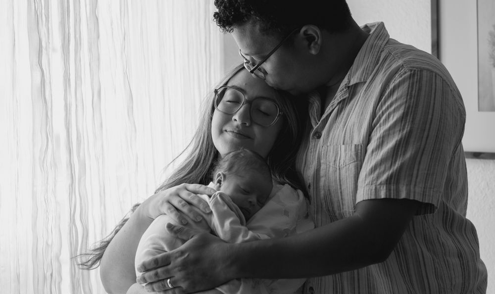Black and white portrait of new parents gently holding and kissing their newborn baby.