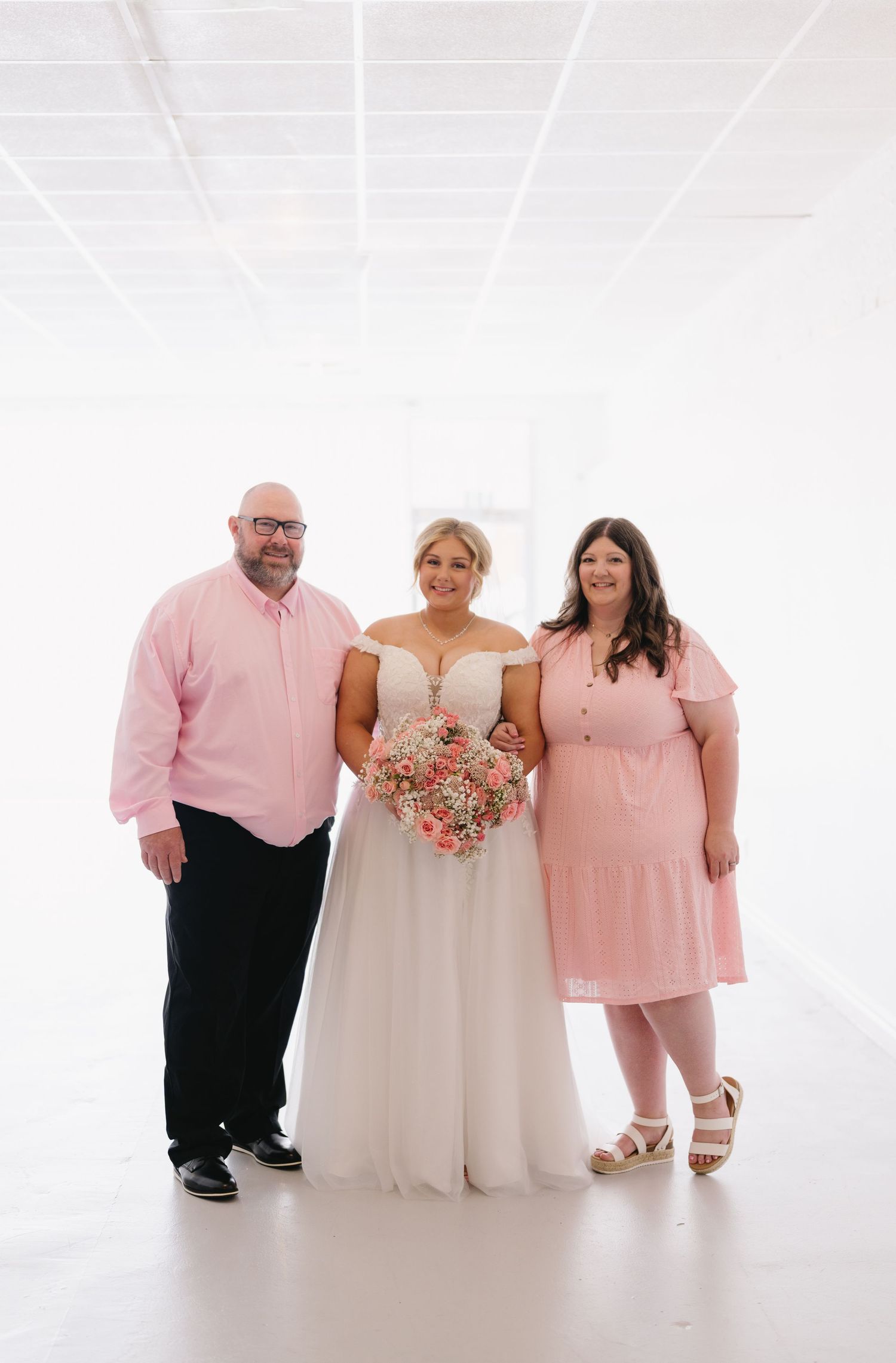 Bride in white off-shoulder gown holds pink bouquet flanked by two guests in pink attire in bright white venue.