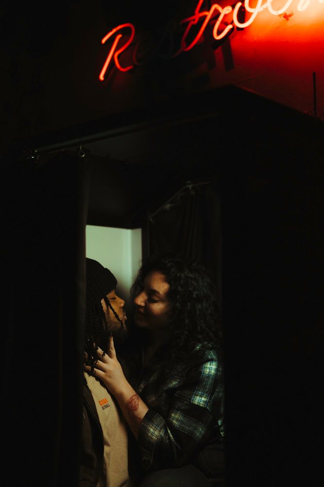 Couple sharing a romantic kiss beneath a red neon Restroom sign in a dark arcade bar.