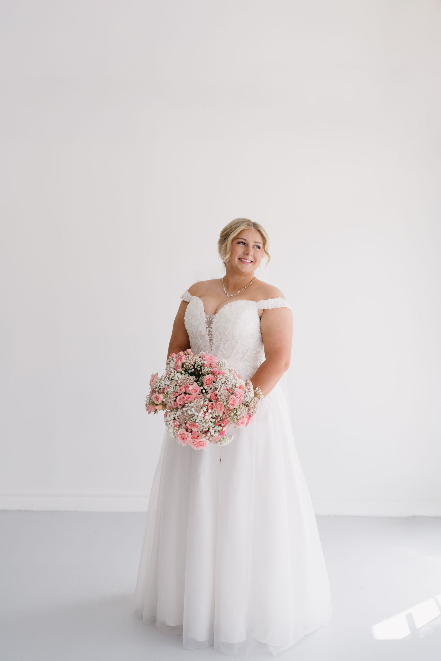 Smiling plus-size bride in off-shoulder lace wedding gown holds pink bouquet against white wall.