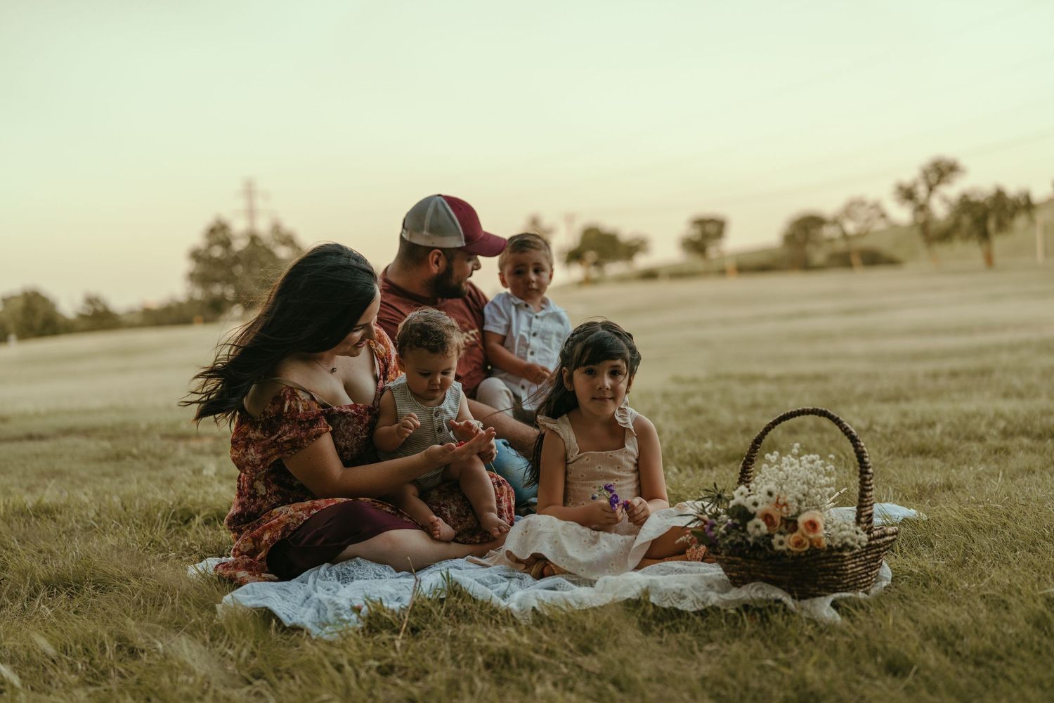 Happy family with three young children enjoying a picnic on a grassy field at golden hour.