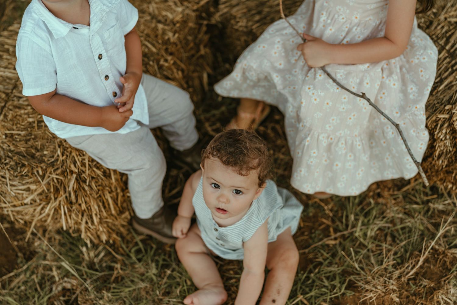 Overhead view of three young children sitting together on golden hay bales outdoors in natural light.