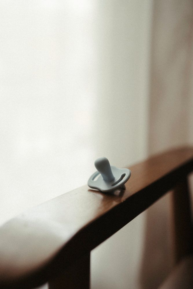 A baby pacifier resting on a wooden crib rail near a soft curtained window.