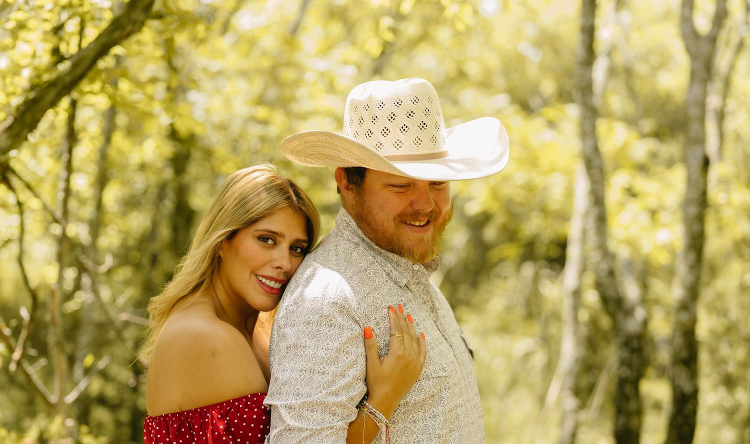 A smiling couple poses outdoors, woman in red dress, man in white cowboy hat and vest, surrounded by green trees.