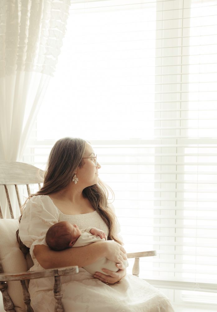 Mother in white dress sitting in rocking chair holding newborn baby by bright window.