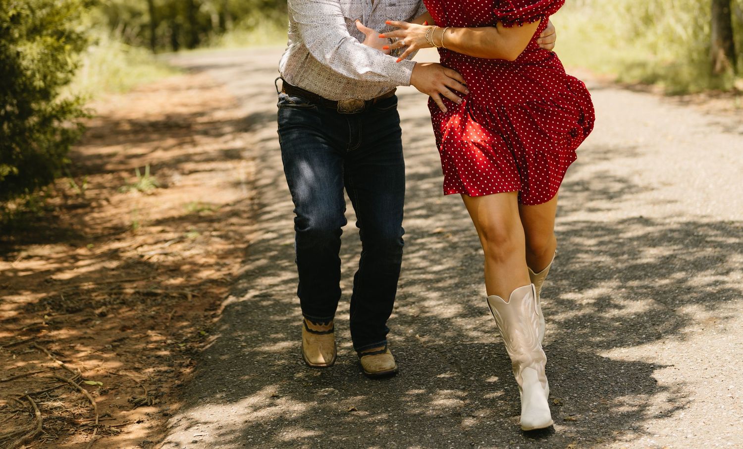 Couple dancing on a dirt path, woman in red polka dot dress and white cowboy boots, man in jeans and boots.