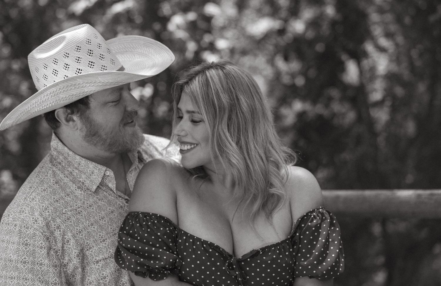 Black and white photo of a smiling couple, man in cowboy hat and woman in polka dot dress, sharing a tender moment outdoors.