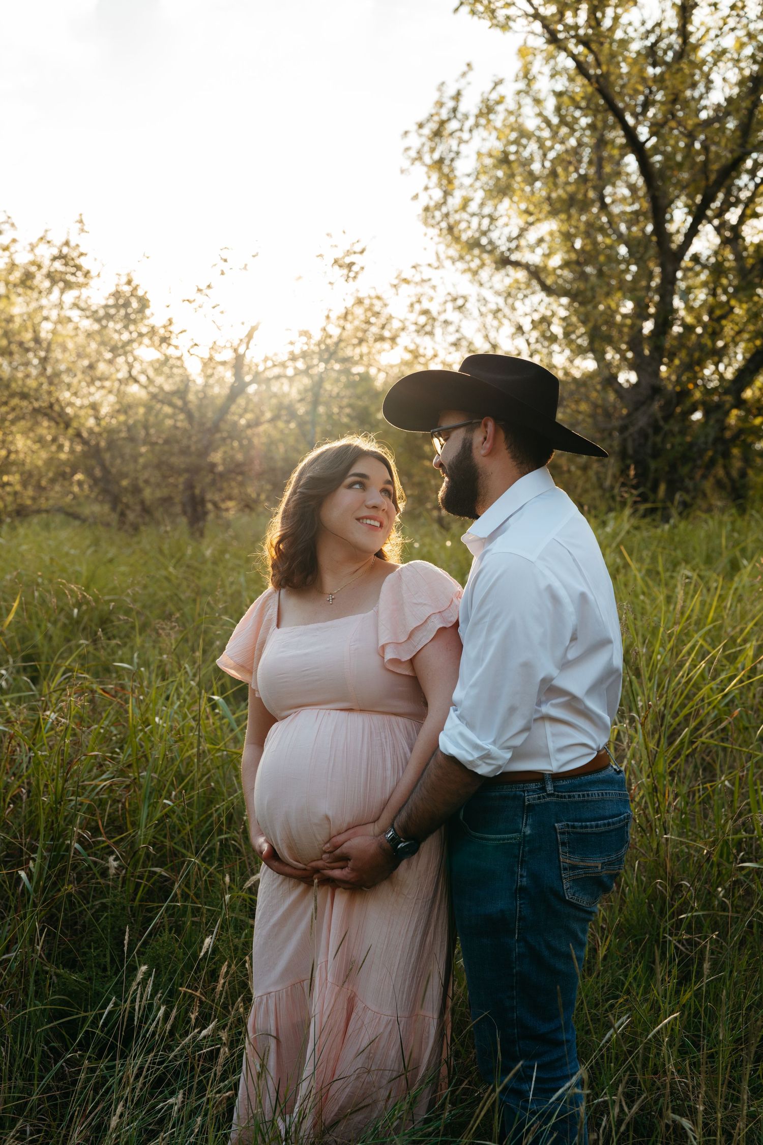 Pregnant couple holding baby bump in golden sunset light in grassy field.