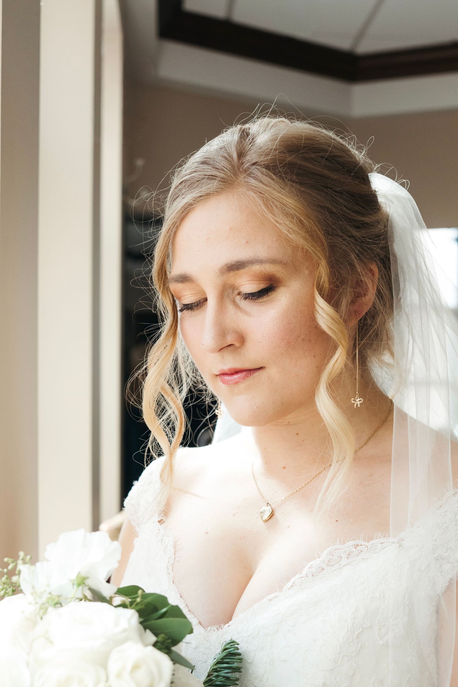 Beautiful blonde bride in white lace gown with veil looks down holding white rose bouquet near window.