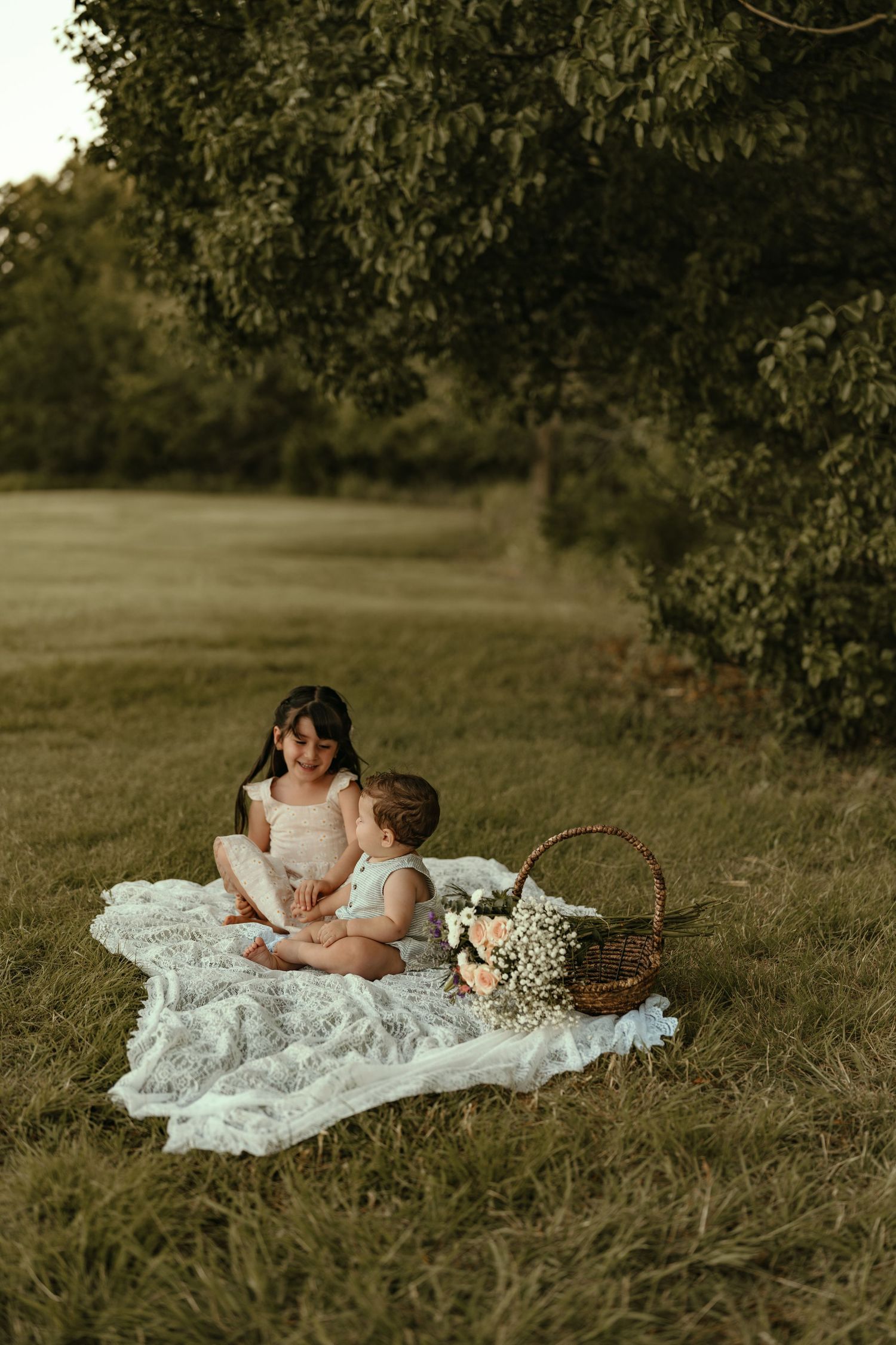 A young girl and baby sit together on a white lace blanket in a sunlit green meadow with flowers.