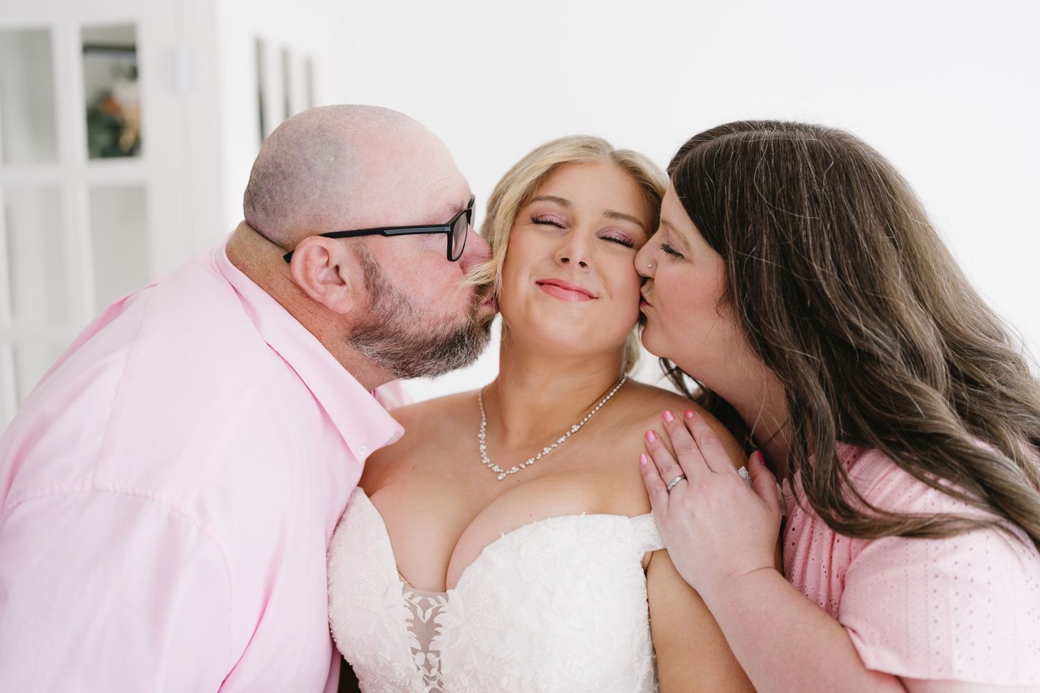 Bald man with glasses and brunette woman in pink kiss bride on cheeks in sweet wedding moment.