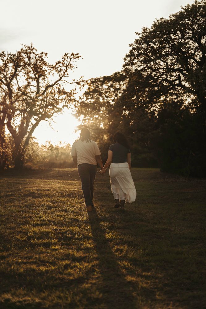 Couple holding hands walking toward a glowing sunset through a lush green meadow with tall trees.