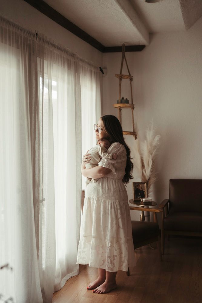 New mother in white lace dress holding newborn baby while gazing out a sunlit window.