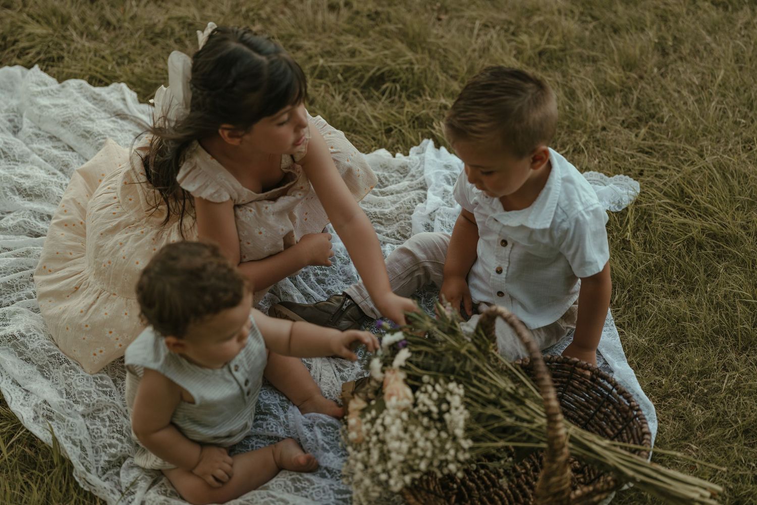 A mother sits on a blanket in a field with her three young children and a flower basket.