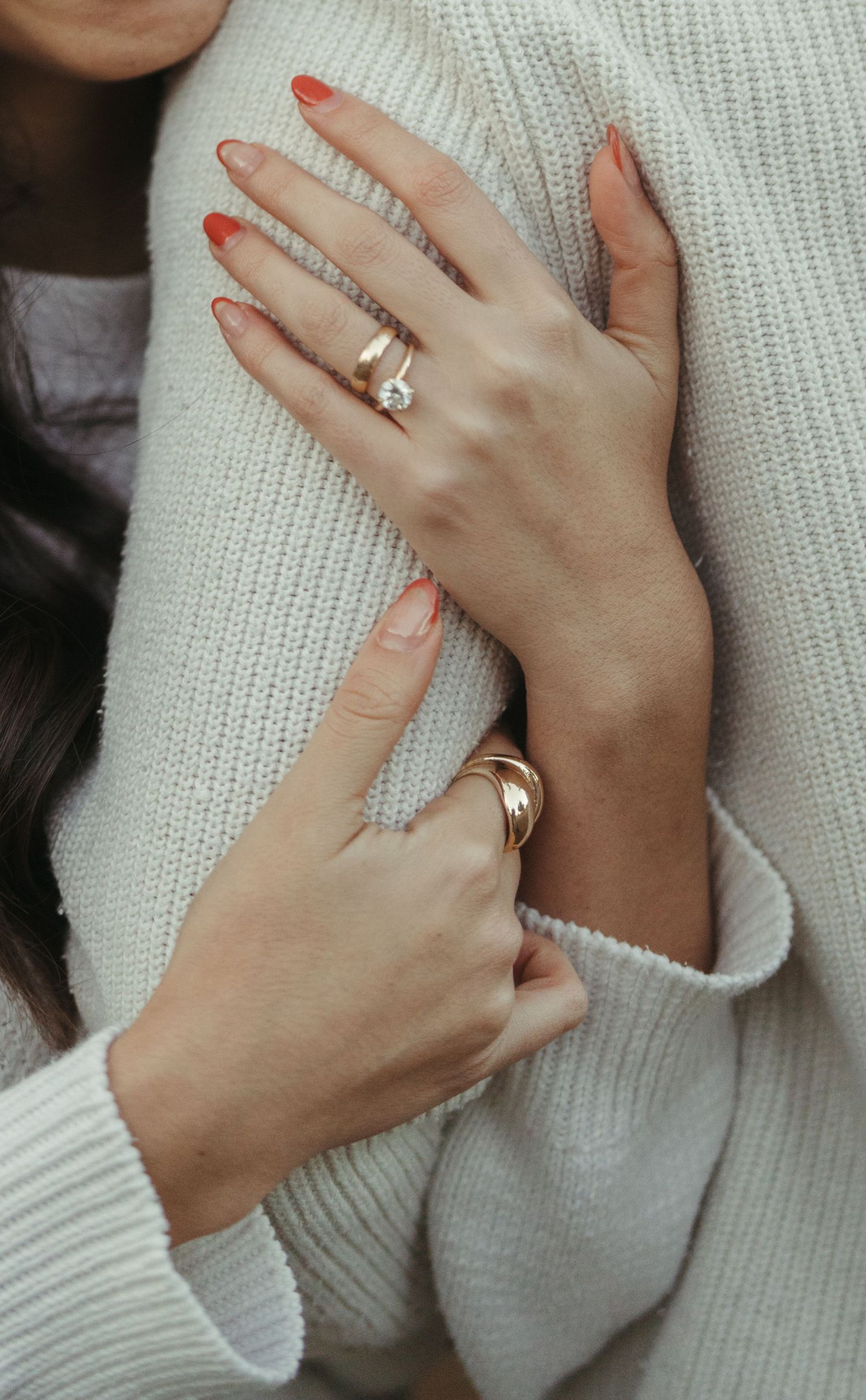 Close up of womans hands with diamond engagement ring and gold band on white sweater.