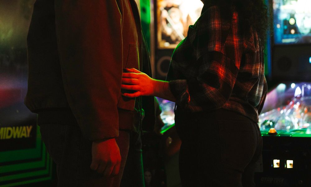 Couple sharing an intimate moment beside a Midway arcade game in a neon-lit arcade bar.