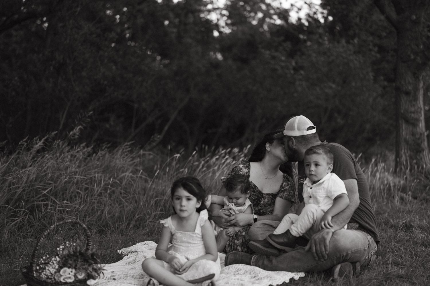 Black and white family portrait of parents with three young children relaxing on a blanket outdoors.