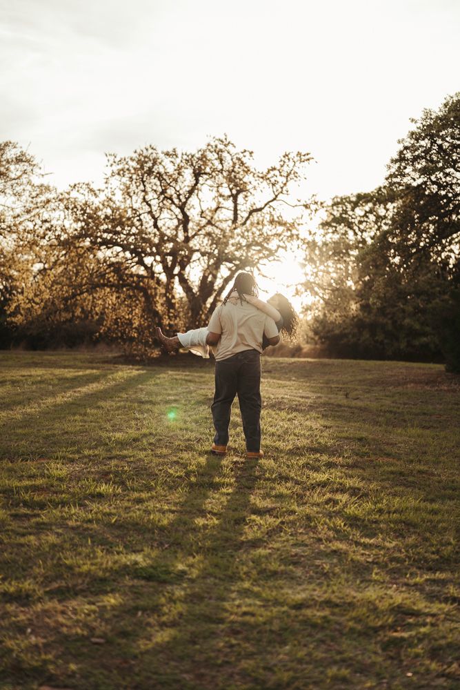 Man carrying a woman in his arms while walking through a sunlit golden hour meadow with trees.