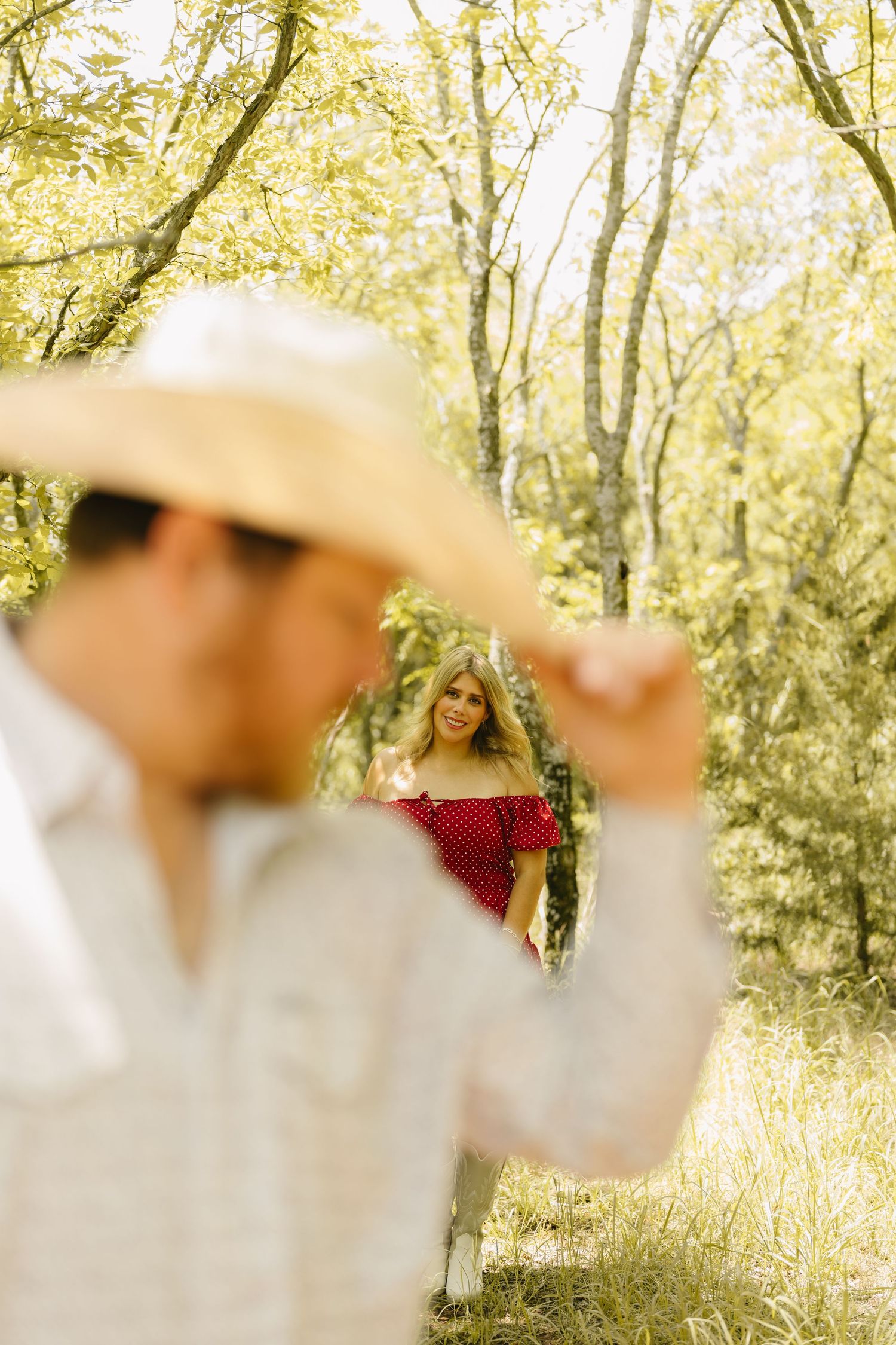 Man in straw cowboy hat with blurred foreground, woman in red off-shoulder top smiling in sunny forest background.