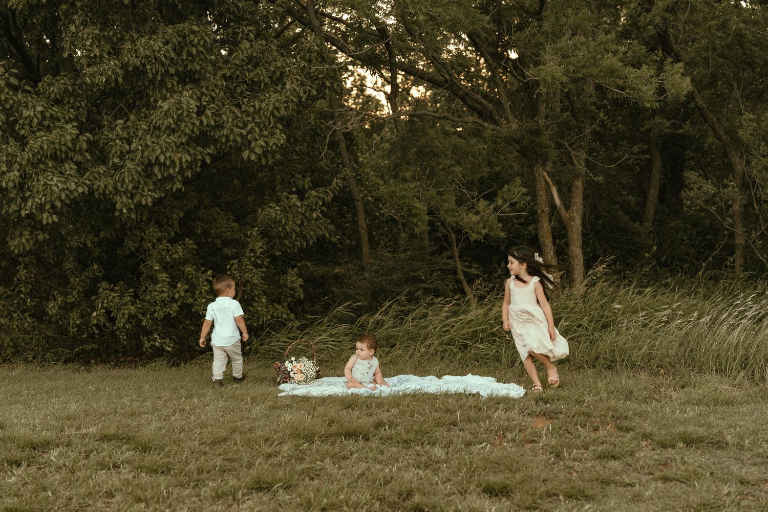 Three young children play freely on a blanket in an open grassy field at dusk.