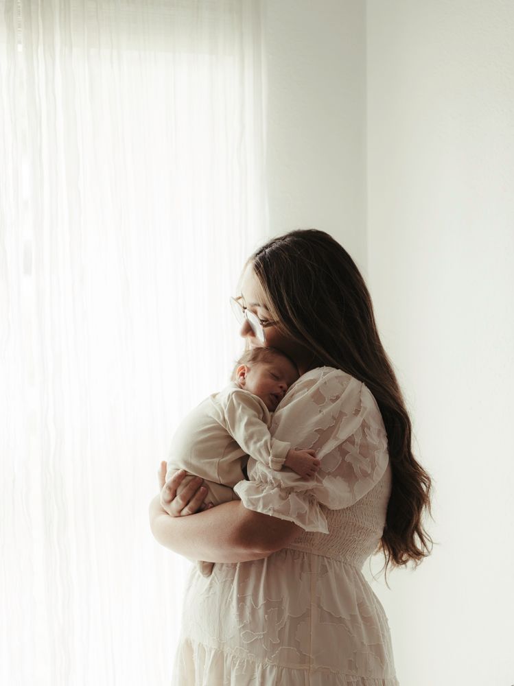 Mother in white lace dress tenderly holding her sleeping newborn baby by a bright window.