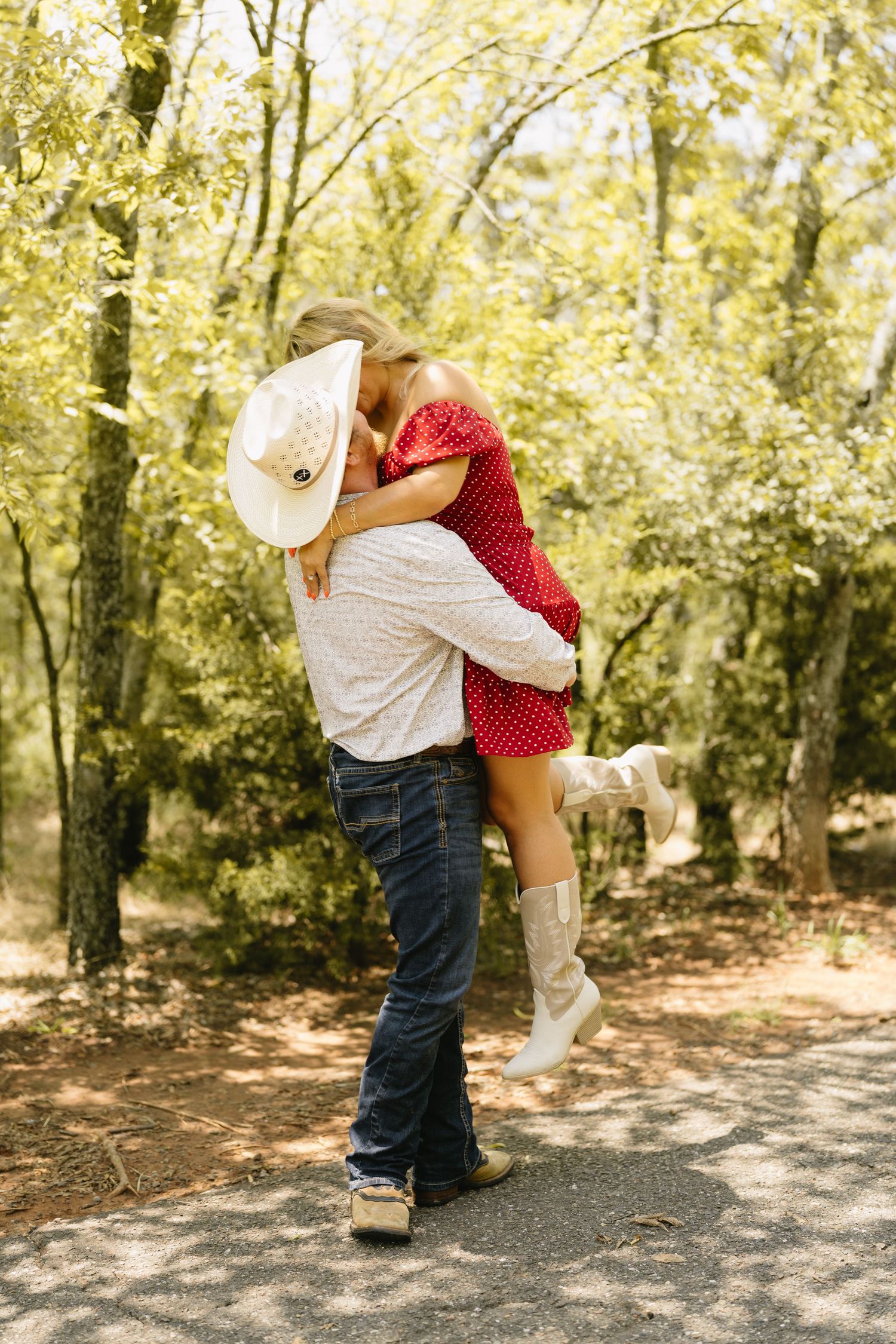Couple sharing a romantic kiss in a sunlit forest, woman in red dress and white boots lifted by man in cowboy hat.
