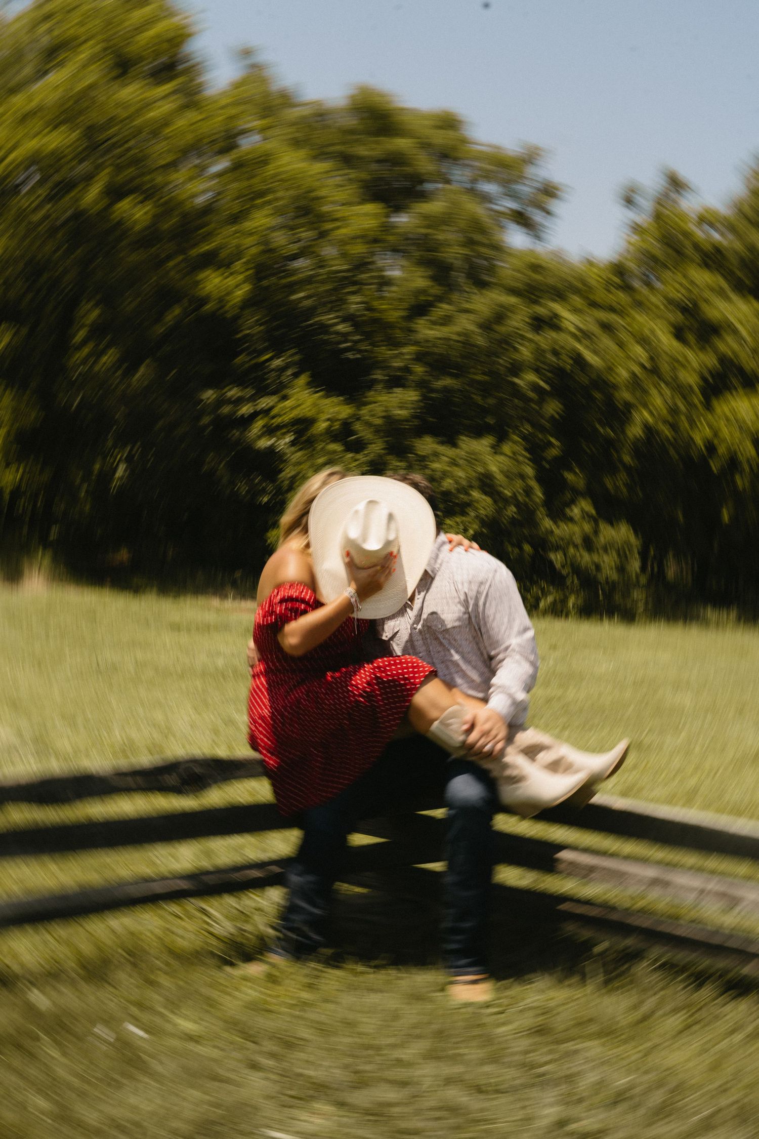 Couple kissing on a rustic fence in a sunny green field, wearing cowboy hats and western attire.